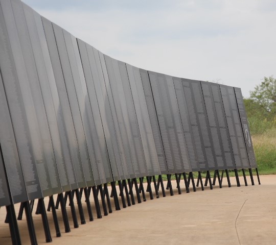 A series of tall, dark metal panels with inscriptions stand in a curved row on a concrete pathway outdoors, with grass and cloudy sky in the background.