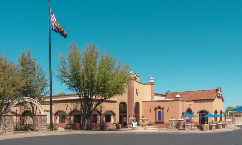 A tan and terracotta building with arched windows and a tiled roof stands under a clear blue sky, shaded by trees. Two flags are raised on a flagpole beside the entrance. Blue umbrellas cover tables on a patio.