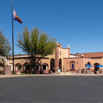 A beige stucco building with arched windows, a central tower, and terra-cotta roof tiles sits under a clear blue sky. An American and Arizona flag fly on a flagpole, and trees provide shade near the entrance.