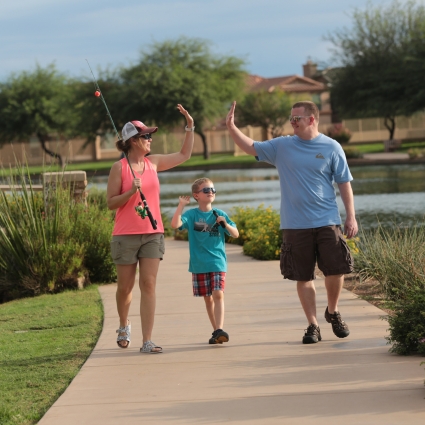 A woman, man, and young boy walk on a path by a pond, smiling and giving each other high-fives. The woman holds a fishing rod. Trees and houses are visible in the background.