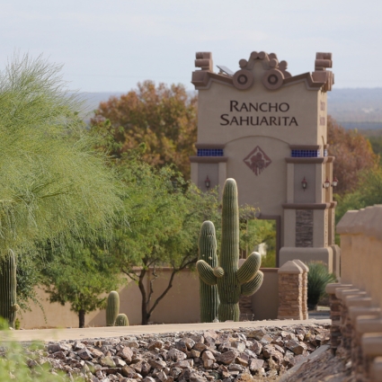 A desert landscape with cacti and rocks in the foreground; in the background, there is a decorative entrance sign that reads "Rancho Sahuarita" surrounded by trees and a stone wall.