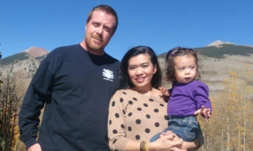 A man, woman, and young child pose outdoors with mountains and trees in the background on a sunny day. The woman holds the child, and all three are looking at the camera and smiling.