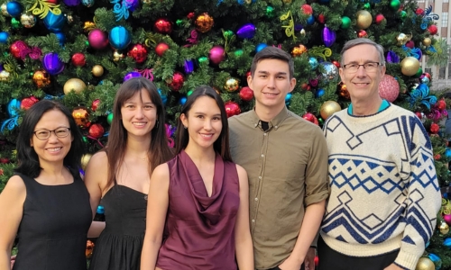 Five people standing in front of a brightly decorated Christmas tree with colorful ornaments, smiling at the camera. The group includes three women and two men dressed in festive, casual clothing.