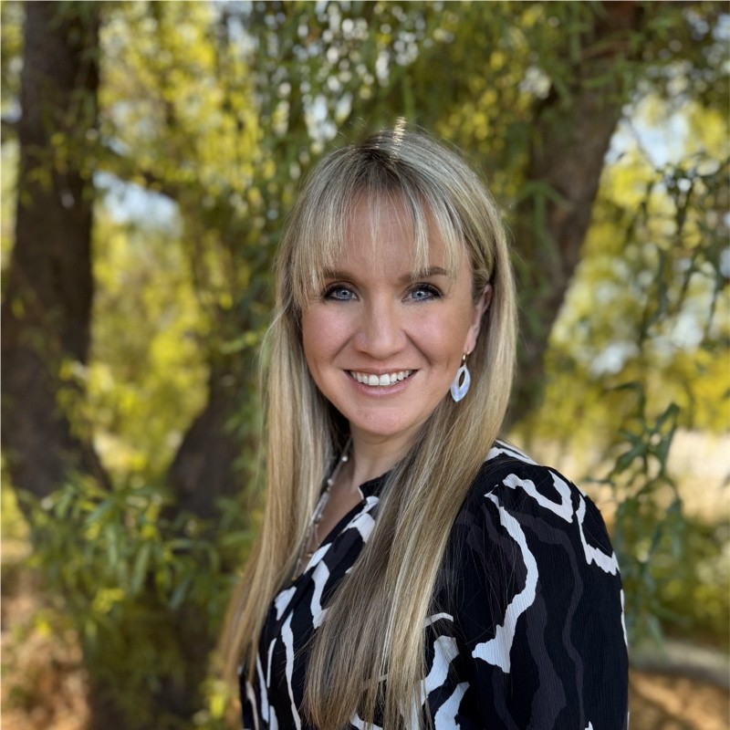 courtney-leon A smiling woman with long blonde hair and blue eyes stands outdoors in front of leafy trees, wearing a black and white patterned blouse and white earrings. Sunlight filters through the branches in the background.