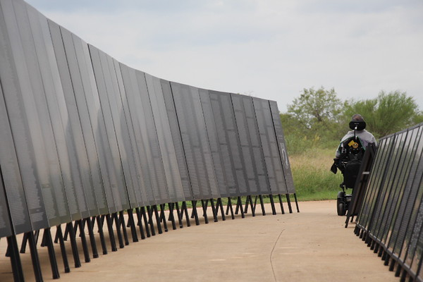 A person in a wheelchair moves along a pathway lined with large, reflective panels that display engraved names, resembling a memorial wall, under an overcast sky with greenery in the background.