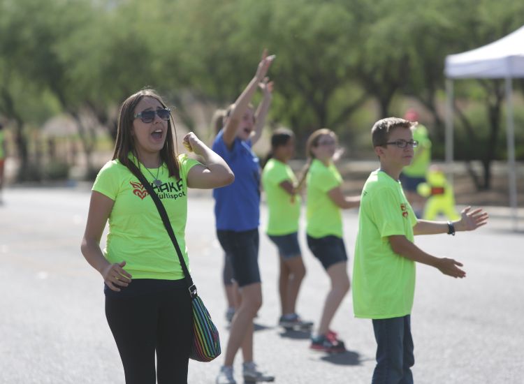 A group of teens wearing bright yellow shirts stand in a line outdoors, cheering and gesturing enthusiastically. One girl in sunglasses is in the foreground, and trees and a white canopy are visible in the background.