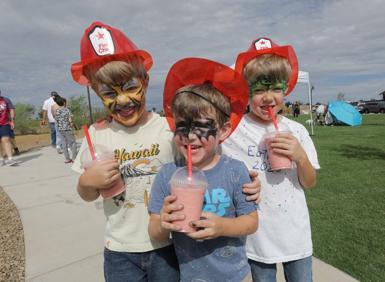 lakesiderun-kids Three young boys wearing red plastic firefighter hats and face paint smile while holding pink drinks with red straws outdoors on a sunny day. There is grass, people, and a blue tent in the background.