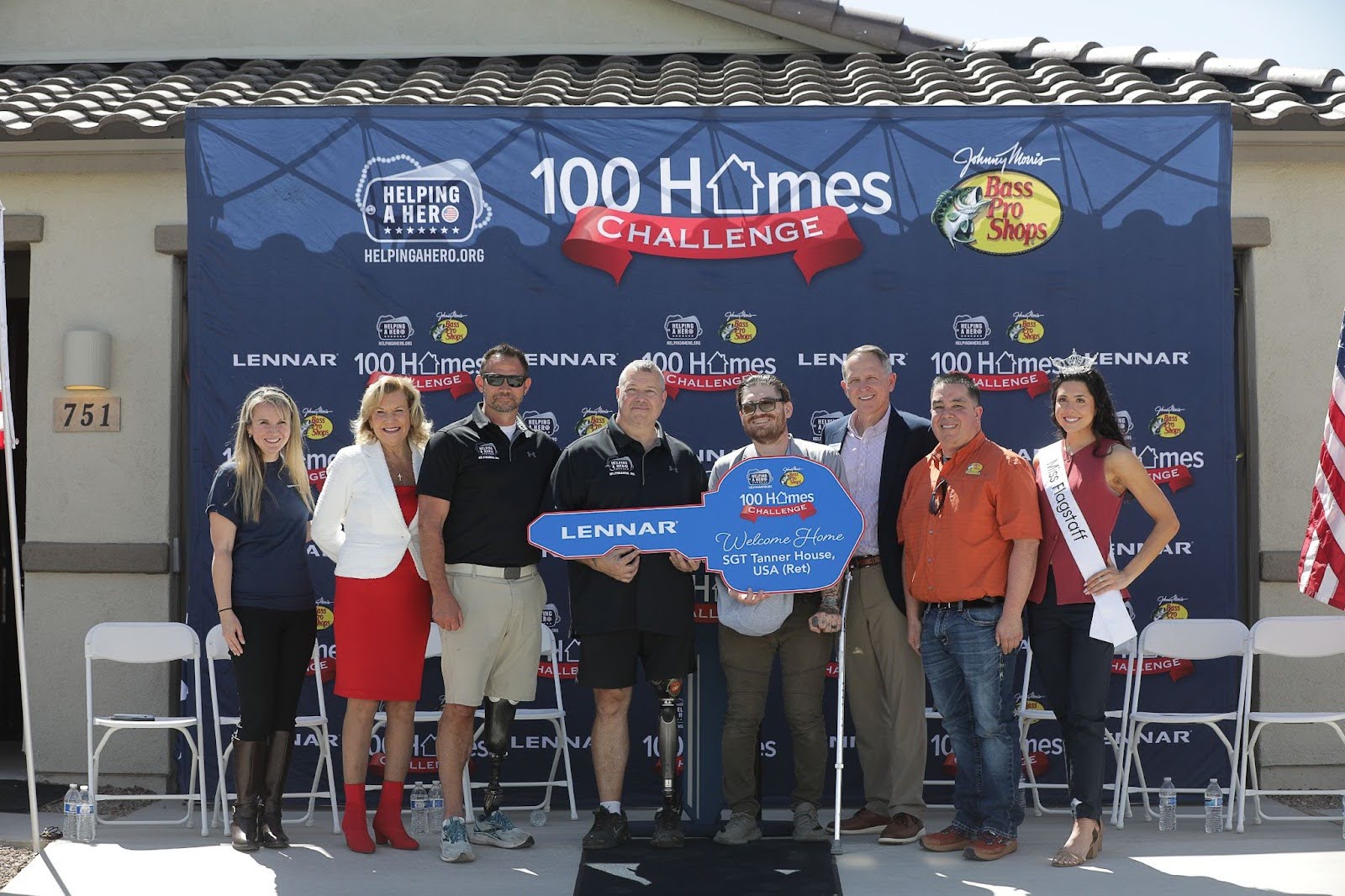 A group of people pose in front of a house holding a large blue ceremonial key at a 100 Homes Challenge event. The backdrop displays sponsors’ logos, and an American flag stands to the right.