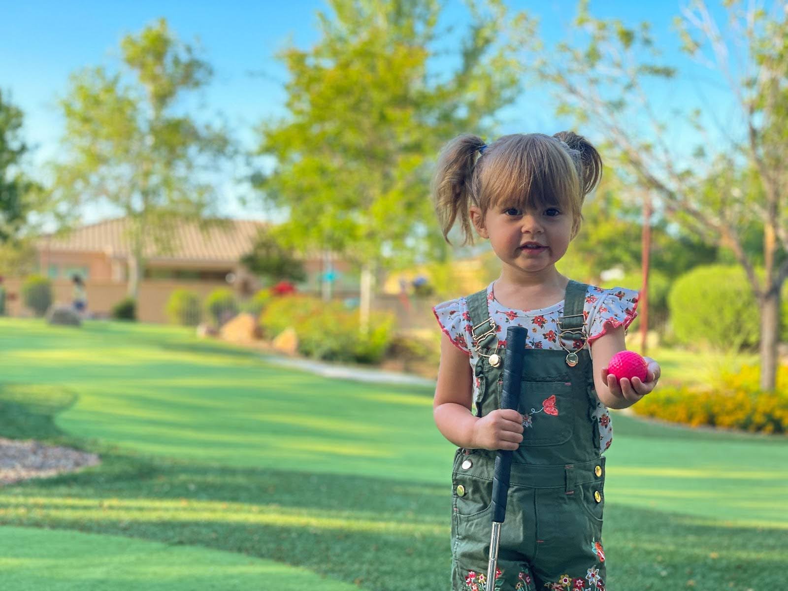 A young girl with pigtails stands on a mini golf course, holding a pink golf ball in one hand and a club in the other, with trees and greenery in the background on a sunny day.