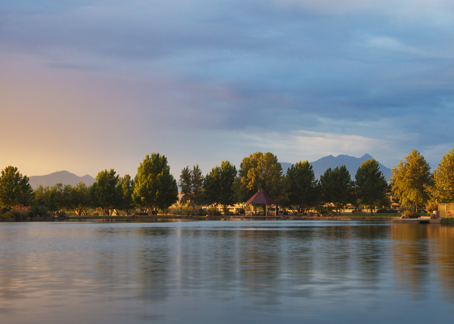 A calm Sahuarita Lake reflects trees and a gazebo at sunset, with mountains visible in the distance under a partly cloudy sky. Warm golden light brightens the scene on the left side.