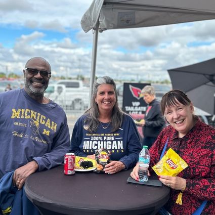 saguaro club Three smiling adults sit at a round outdoor table with snacks and drinks. Two wear Michigan shirts. The sky is partly cloudy, and a sports stadium is visible in the background.