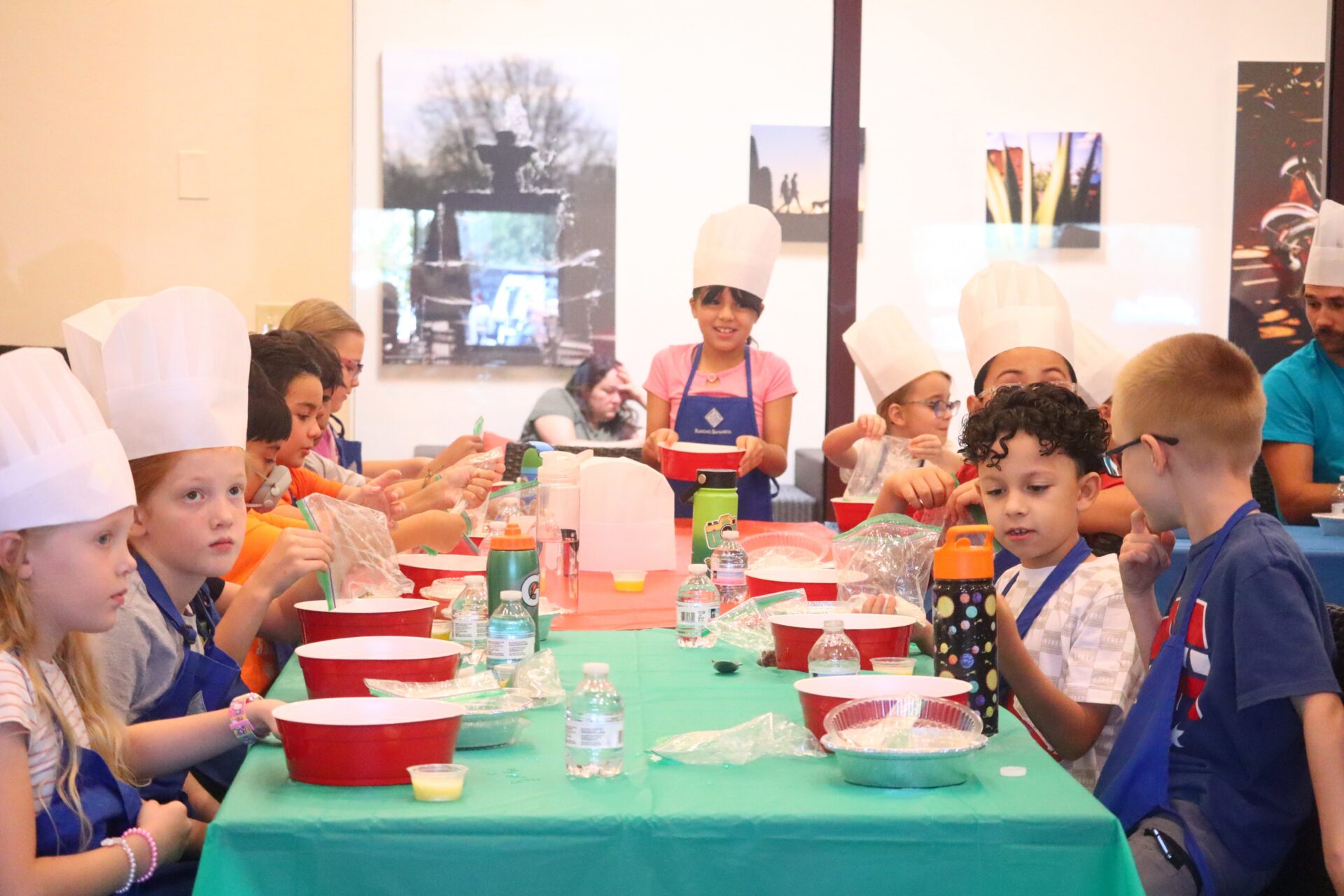 A group of children wearing chef hats and aprons sit around tables with bowls and utensils, participating in a cooking activity. An adult is present in the background. The atmosphere is lively and collaborative.