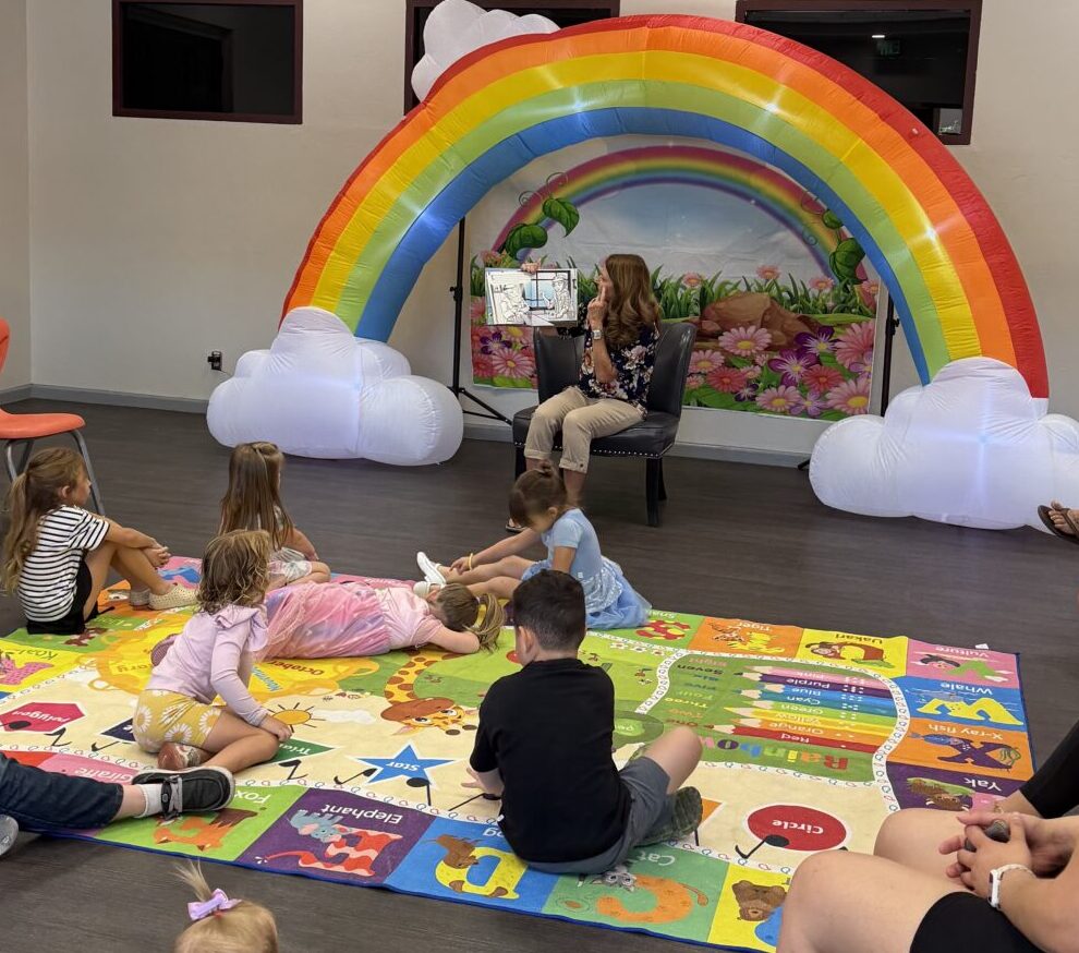 A woman reads a picture book to a group of children sitting on a colorful play mat, in front of a large inflatable rainbow with cloud ends inside a bright room. Adults sit nearby, watching the storytime.