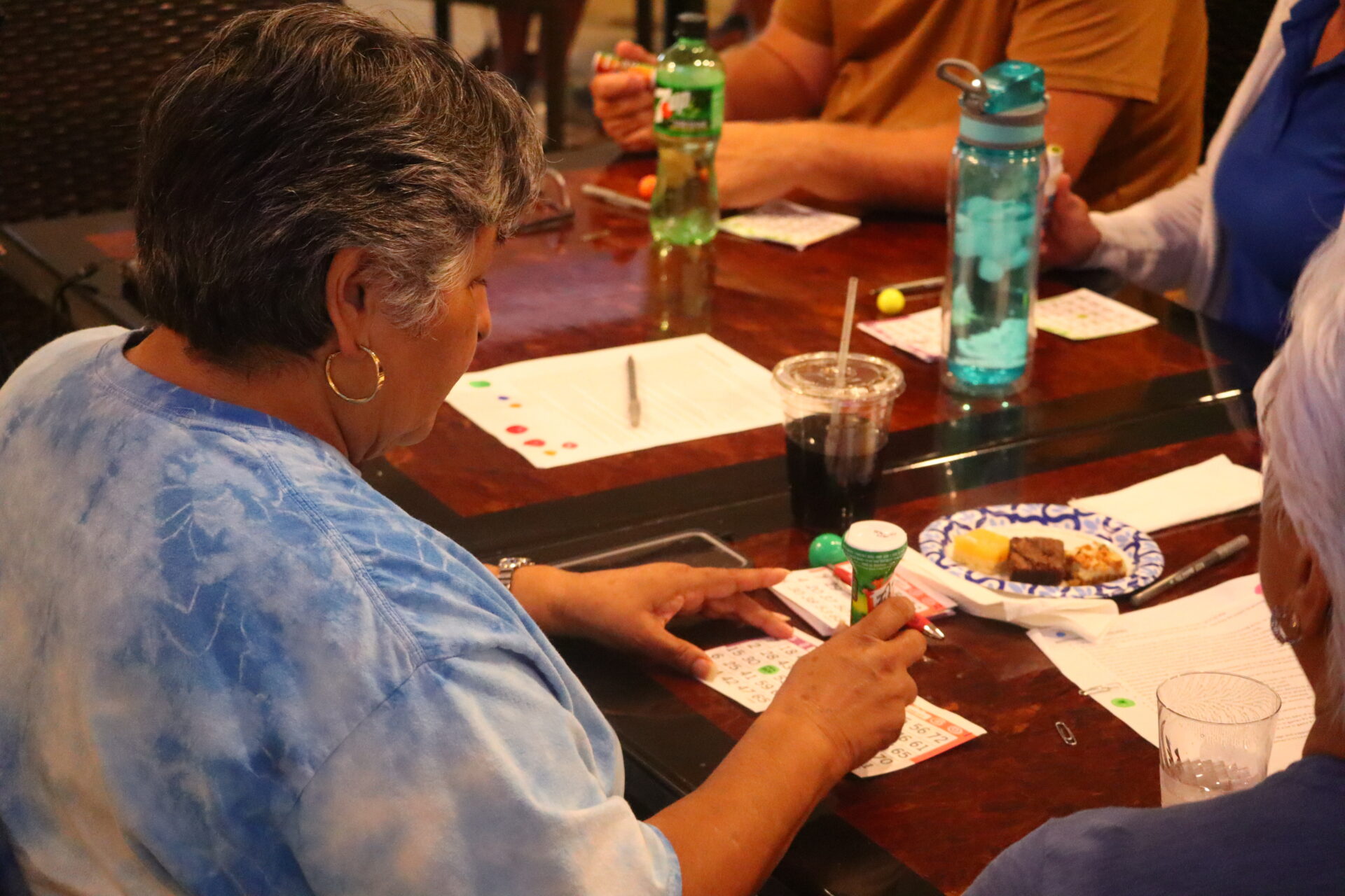 People playing bingo around a wooden table with drinks and snacks.