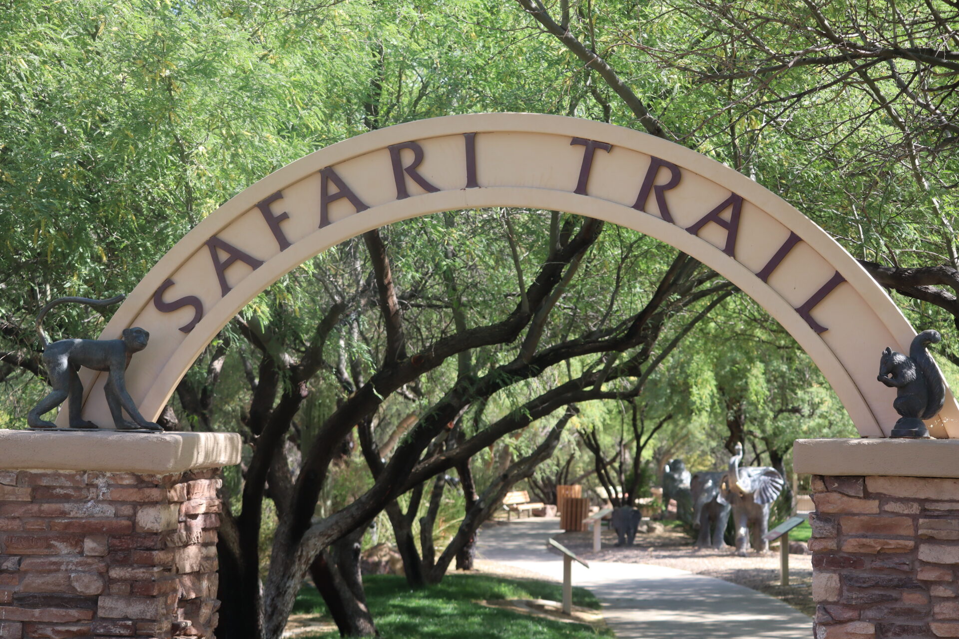 An arched entrance sign reading "Safari Trail" sits above a pathway surrounded by green trees, with animal statues such as elephants visible along the trail. Two small animal statues sit atop the entrance pillars.