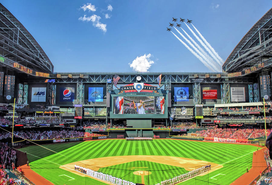 A baseball stadium filled with fans, players lined up on the field, and five jets flying in formation overhead, leaving white trails in the blue sky; a large screen displays a patriotic image.