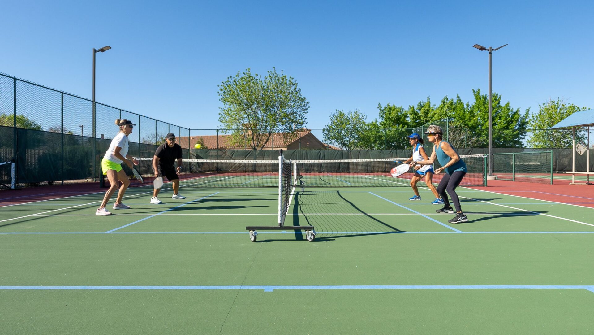 Four people are playing doubles pickleball on an outdoor court under a clear blue sky, with trees and a building in the background—all in your backyard community, enjoying friendly matches with neighbors.
