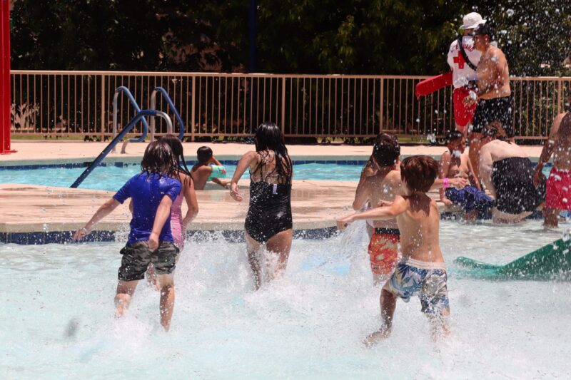 Children run and splash into a swimming pool on a sunny day, while a lifeguard and other people watch and play near the pool’s edge. Trees and a fence are visible in the background.