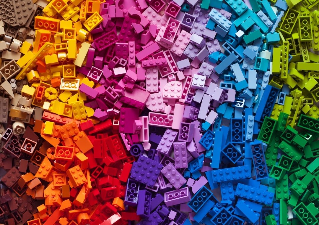 A large assortment of LEGO bricks arranged by color in a gradient, transitioning smoothly from brown and yellow on the left, through orange, red, pink, purple, blue, and finally to green on the right.