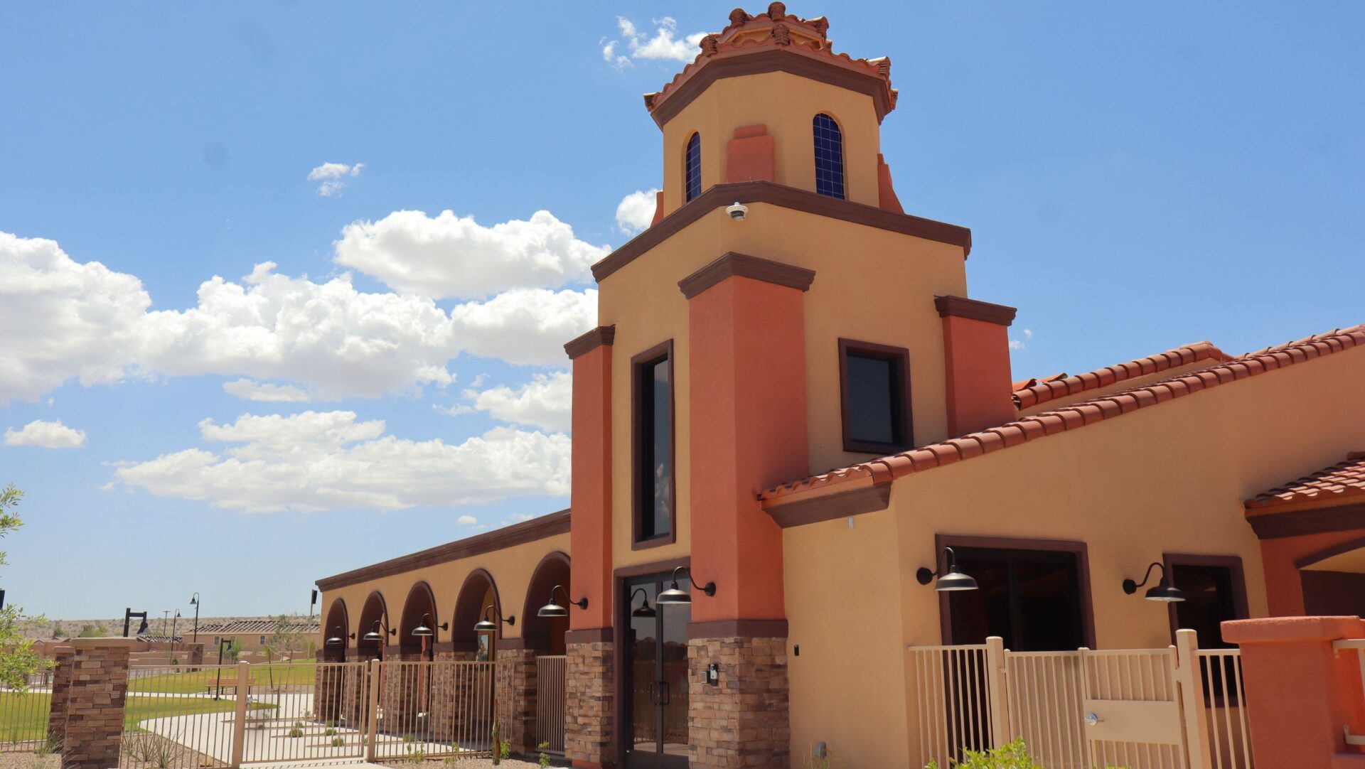 Entrada Del Toro A Spanish-style building with an orange stucco exterior, arched doorways, a tall tower, red tile roof, and a fenced patio, under a bright blue sky with scattered white clouds—an inviting vision reminiscent of the new homes Rancho Sahuarita 2026 will showcase.