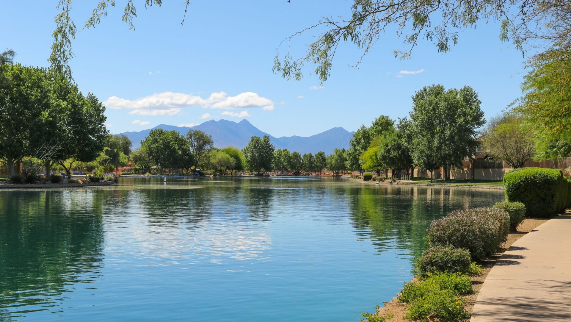 A calm lake bordered by trees, shrubs, and a walking path, with distant mountains under a clear blue sky.