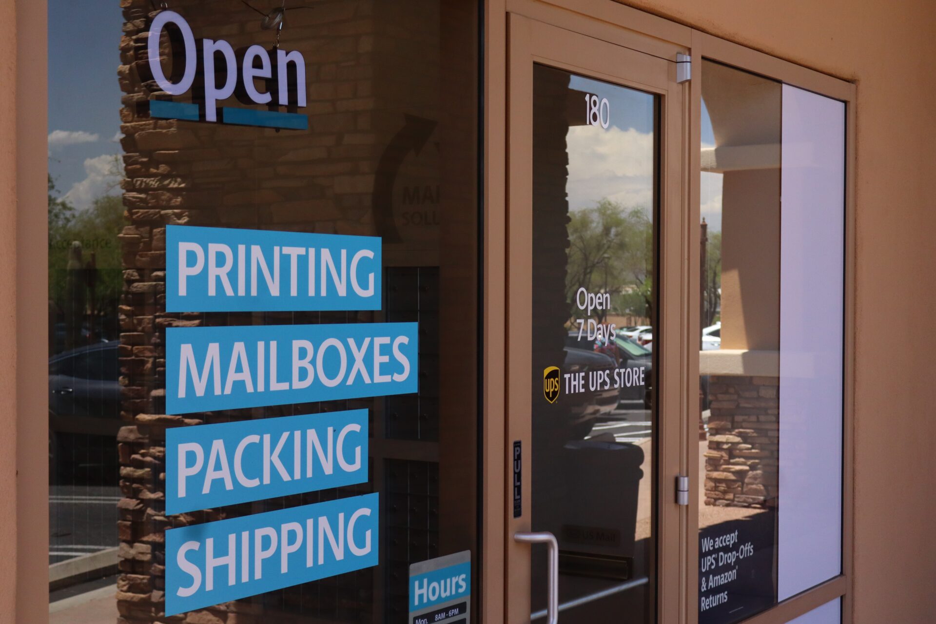 A storefront with signs reading "Open," "Printing," "Mailboxes," "Packing," and "Shipping" in blue and white text. The UPS Store logo and hours are visible on the glass door.
