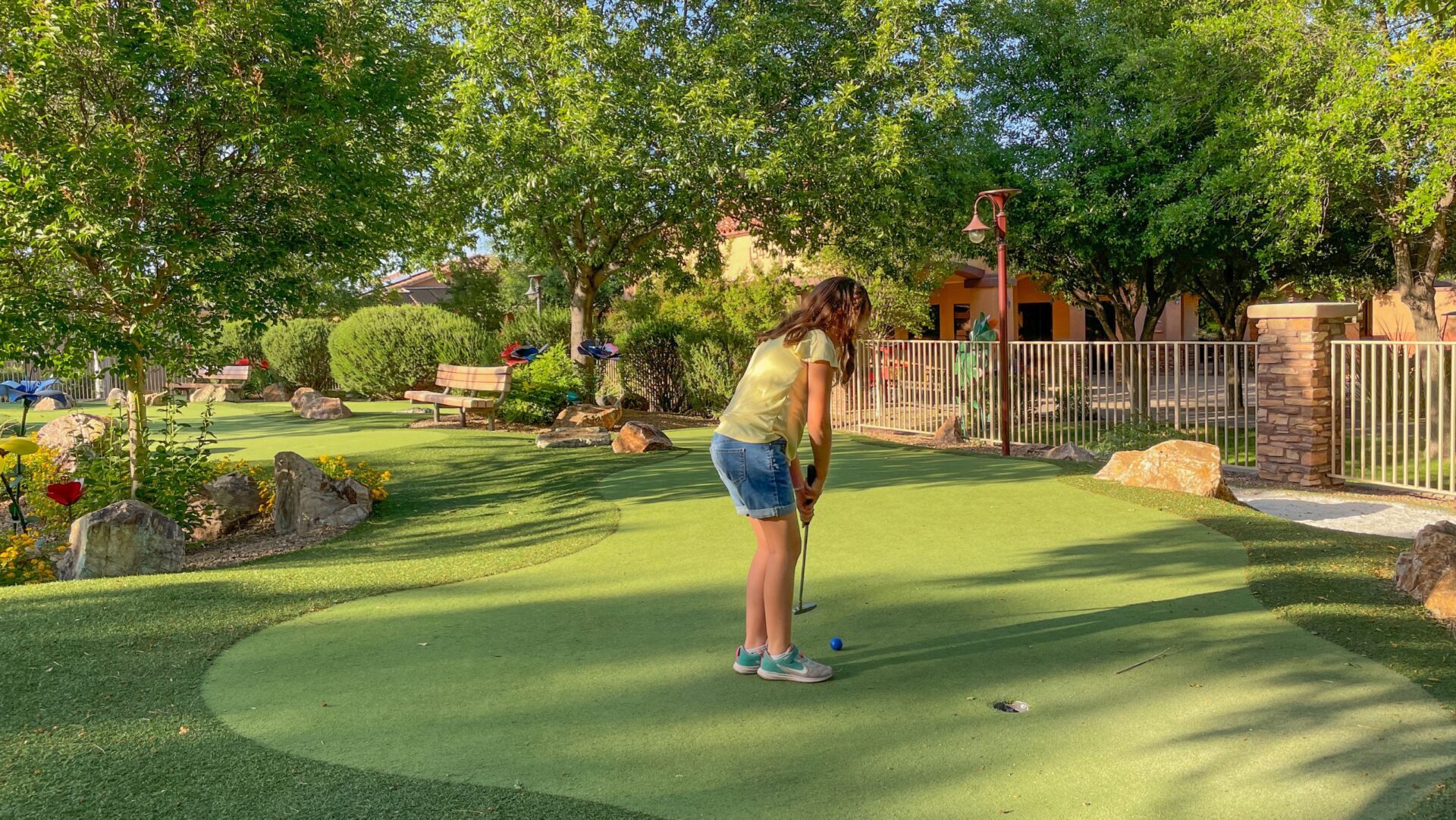 A girl in a yellow shirt and denim shorts plays mini-golf on a sunny day, preparing to putt the ball into the hole on a green, tree-lined course.