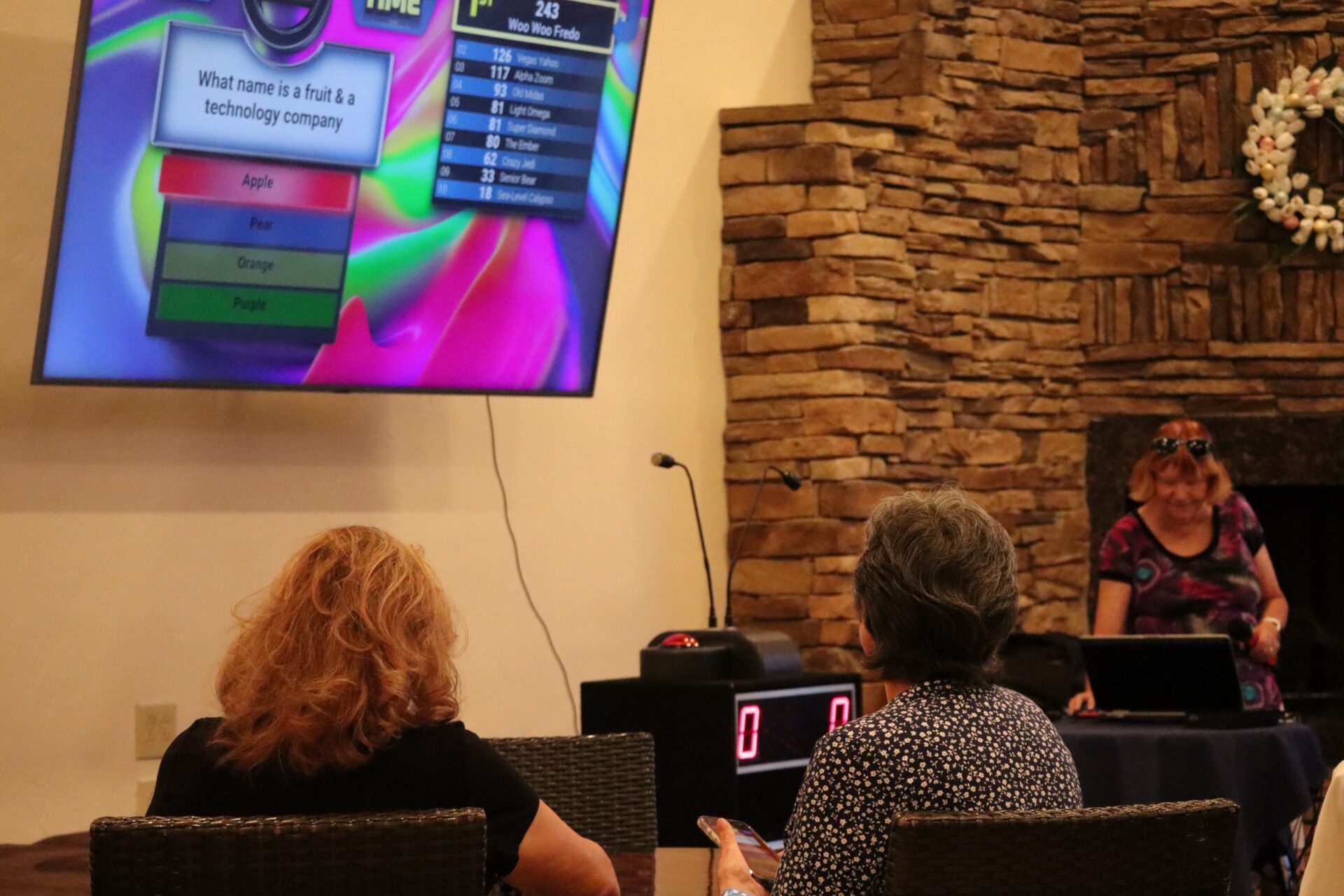 Two women sit facing a large screen showing a trivia question about a fruit and technology company, with answer choices including Apple, while a woman stands at a table with equipment in front of a stone fireplace.