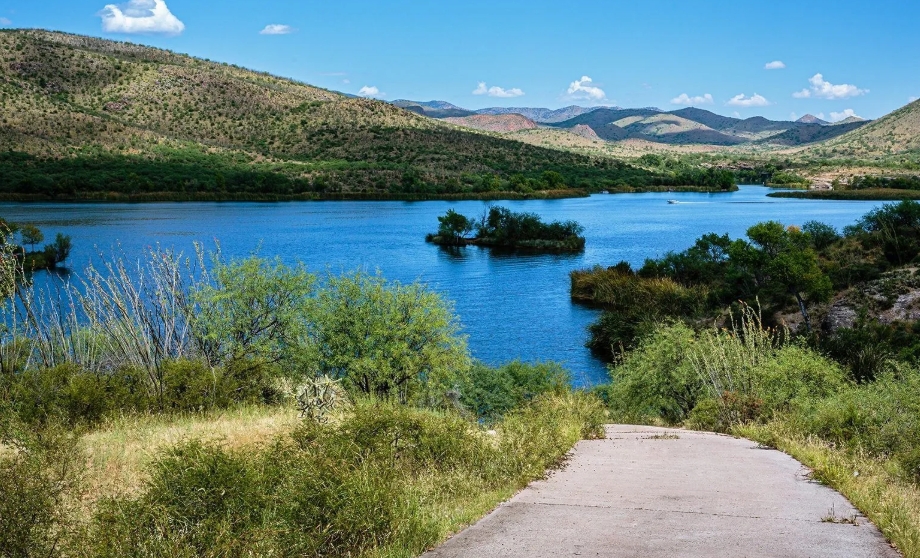 A concrete path leads to a blue lake surrounded by green hills and scattered bushes under a bright blue sky with a few clouds.