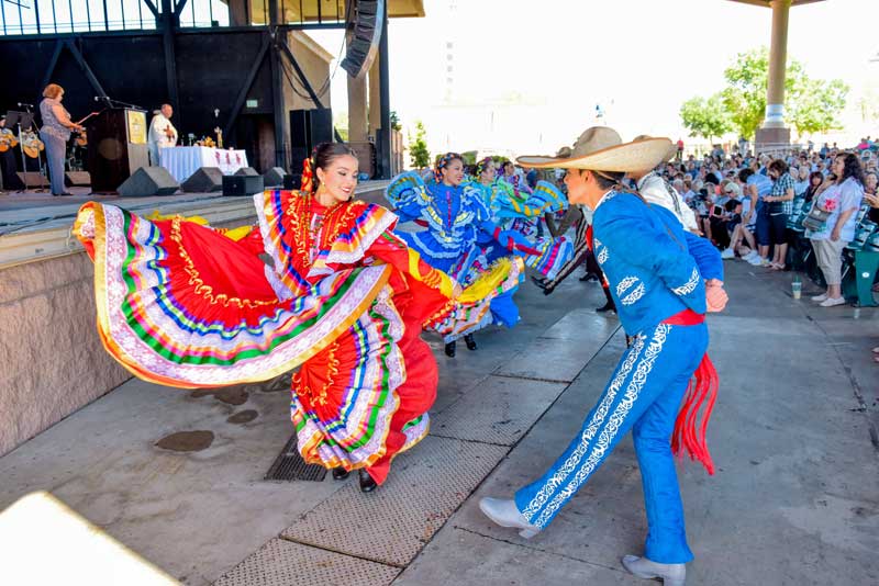 A female dancer in a colorful folk dress and a male dancer in a blue charro suit perform on a stage before an audience.