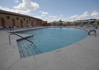 A large outdoor swimming pool surrounded by a concrete deck with rows of lounge chairs and blue umbrellas.