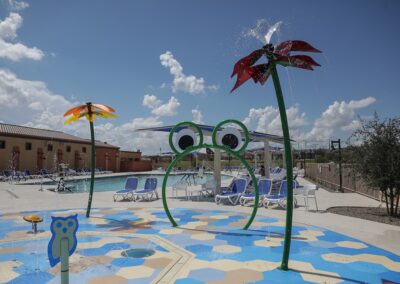 A splash pad with frog-shaped and palm tree-shaped water features in front of an outdoor swimming pool area with lounge chairs.