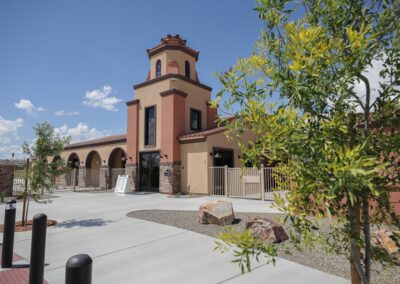 Exterior view of a tan and terracotta-colored building with a central tower and arched walkways under a clear blue sky.