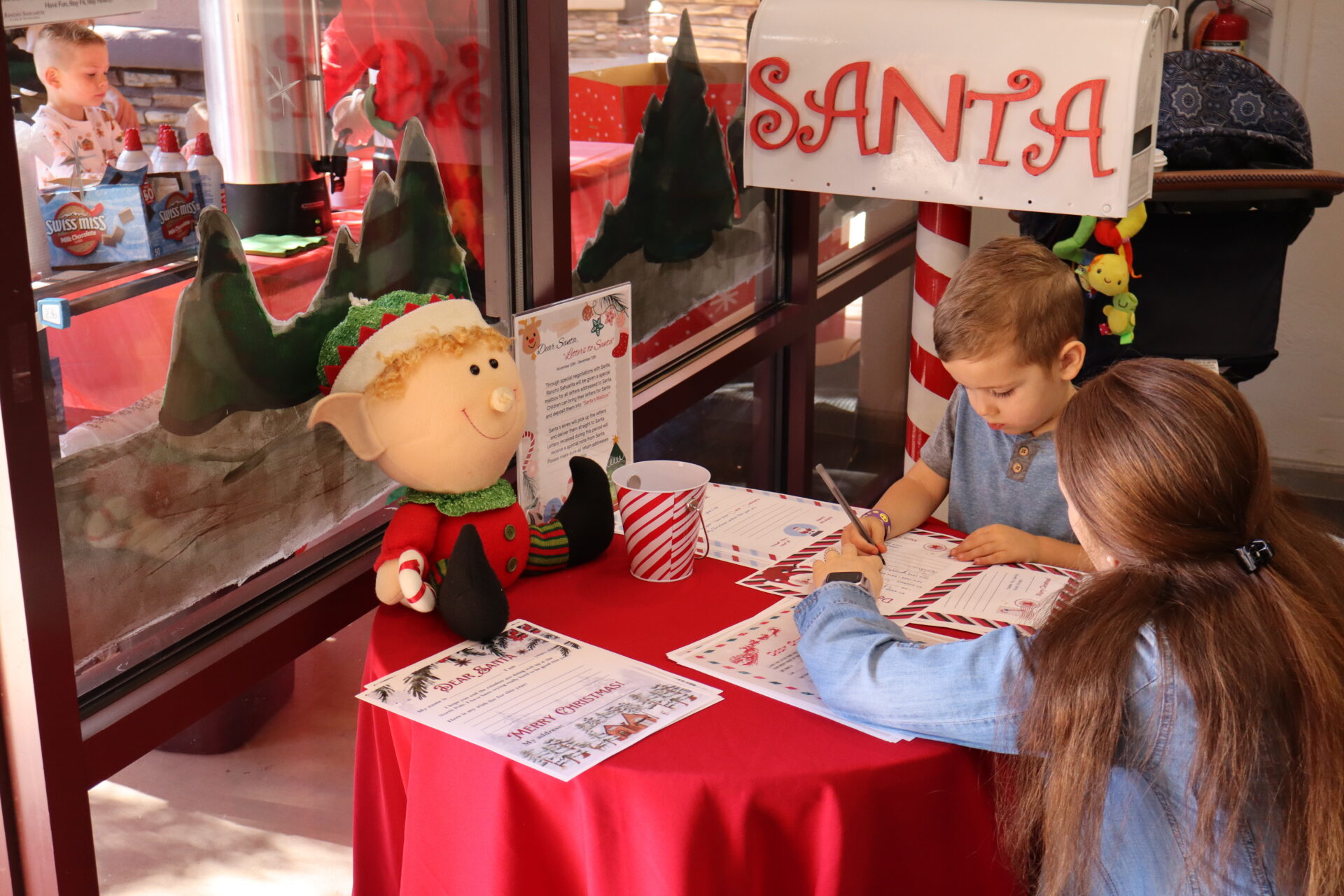 A young boy and girl sit at a small red table, writing letters to Santa next to a plush elf doll.