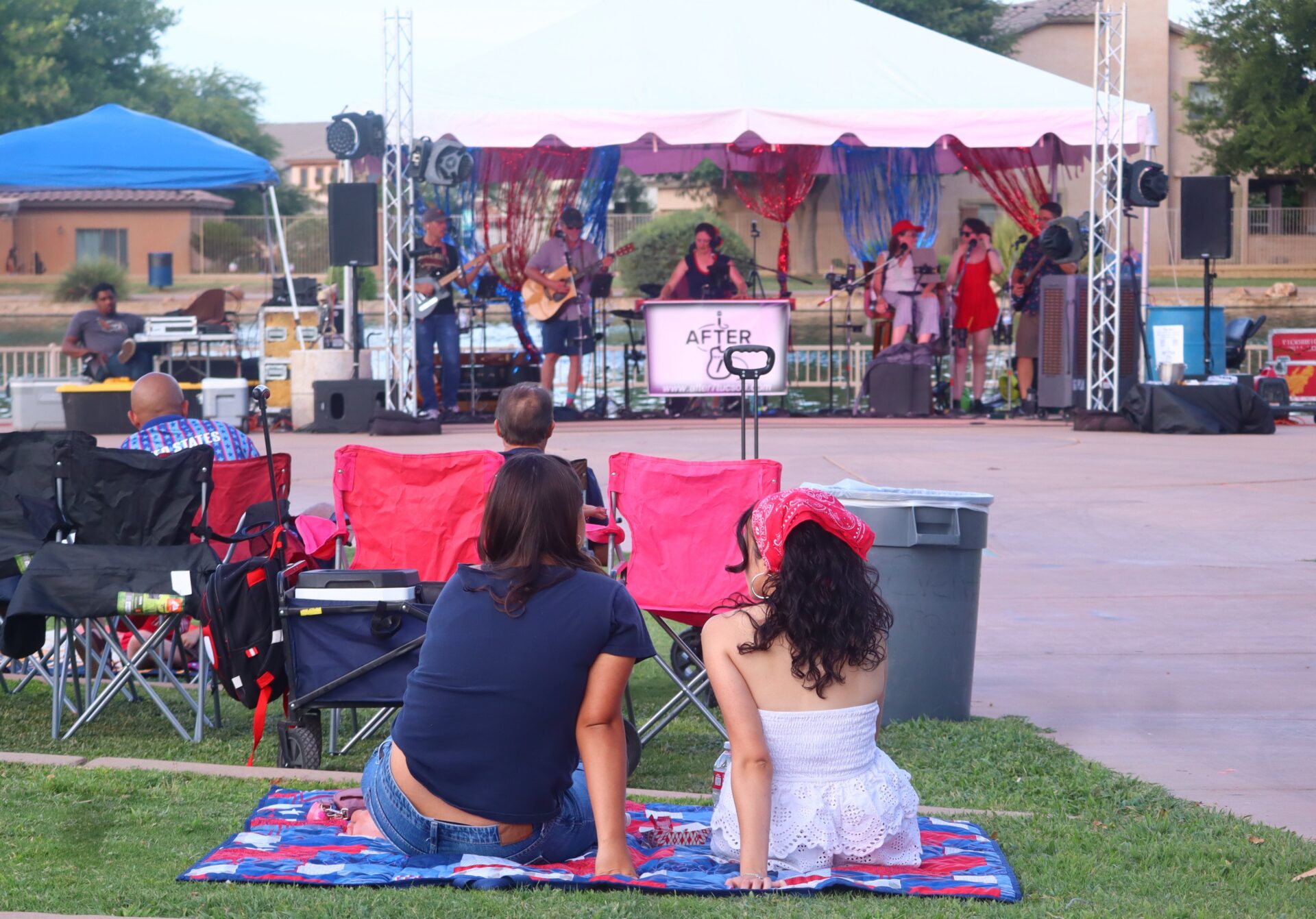 Two women sit on a blanket in a park, watching a live band perform on a stage under a white canopy.