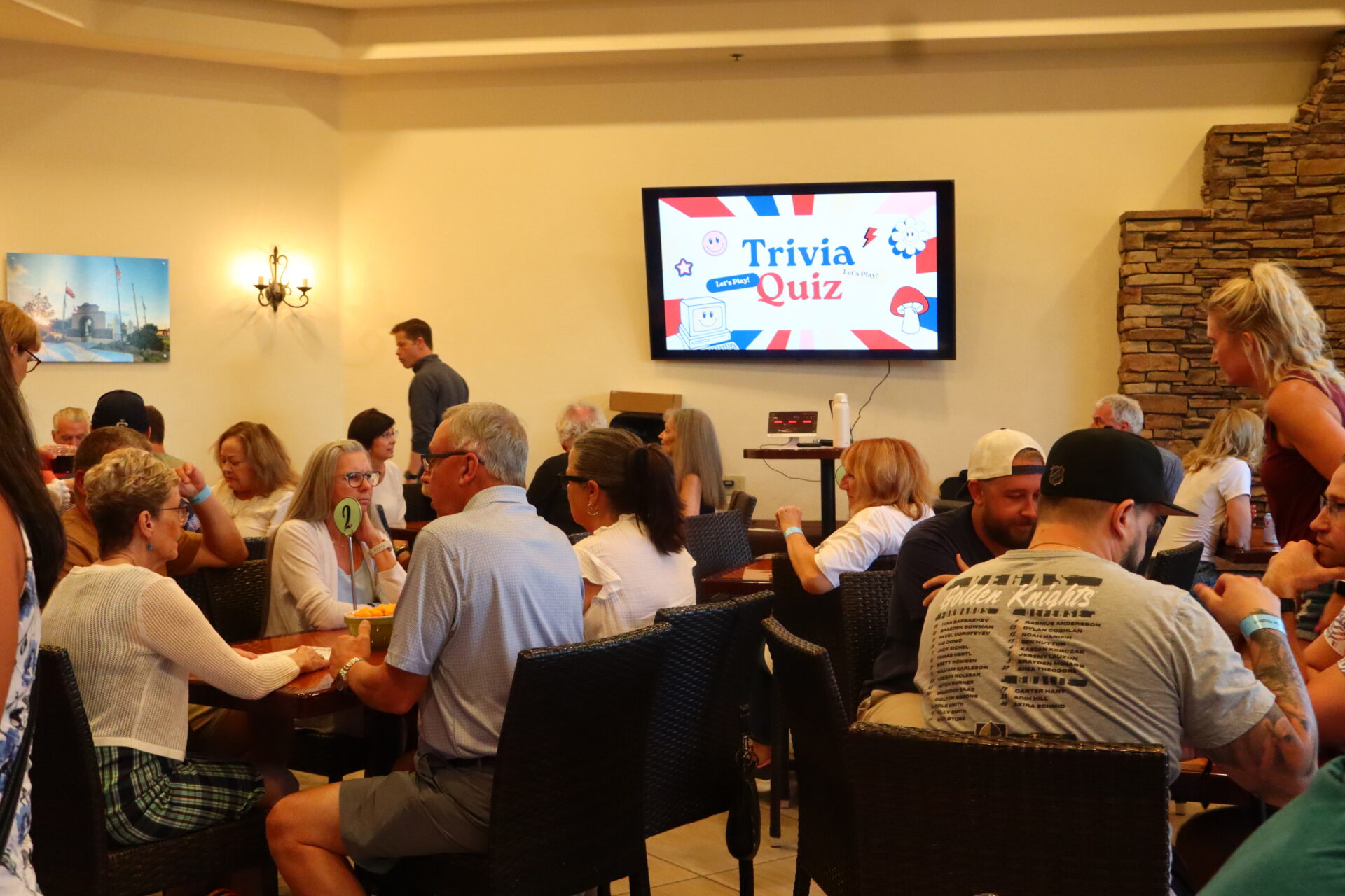 People sitting at tables in a room with a television screen displaying a “Trivia Quiz” graphic.