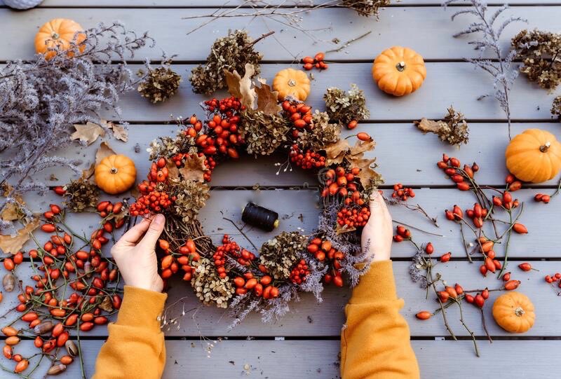 Hands in yellow sleeves arrange a wreath made of dried flowers and rose hips on a wooden table scattered with small pumpkins and berry sprigs.